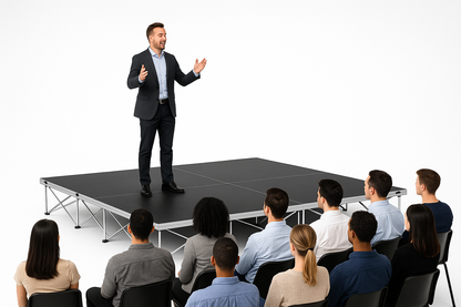 Man giving a presentation to an audience on a LimeLight Lite Series 12x12 modular stage platform in Black HexCoat Laminate at 16" height. Ideal for schools, churches, and event professionals.