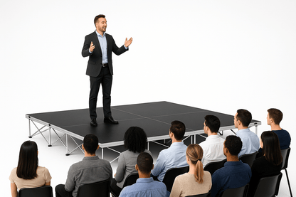 Man giving a presentation to an audience on a LimeLight Lite Series 12x12 modular stage platform in Black HexCoat Laminate at 16" height. Ideal for schools, churches, and event professionals.