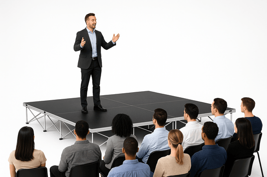 Man giving a presentation to an audience on a LimeLight Lite Series 12x12 modular stage platform in Black HexCoat Laminate at 16" height. Ideal for schools, churches, and event professionals.